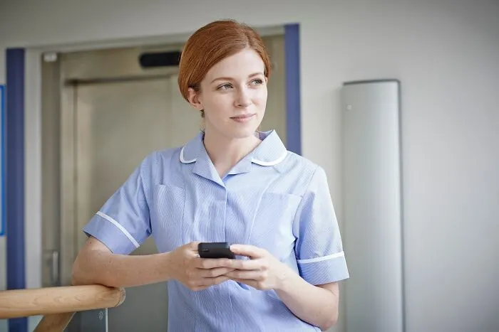 Young female coworker in blue uniform holding smartphone, looking away thoughtfully in a modern workplace setting.