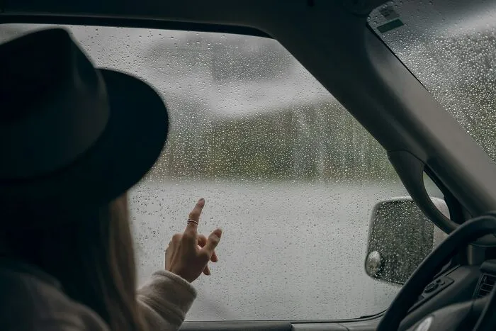 Person wearing a hat sitting in a car, pointing at raindrops on the window, capturing a thoughtful pause moment.