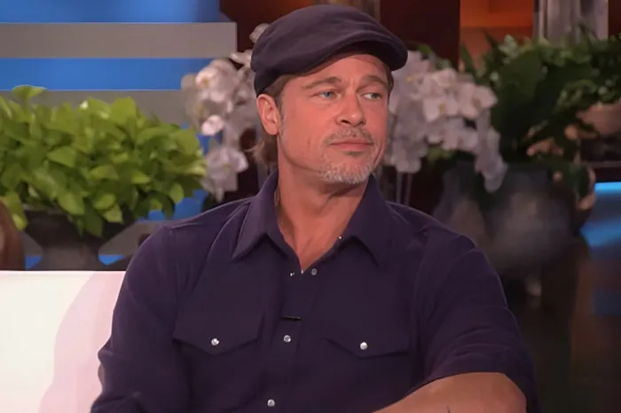 Brad Pitt wearing a navy shirt and cap during a talk show interview, surrounded by plants and studio decor.