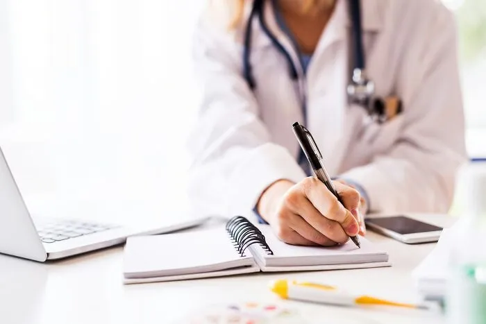 Doctor wearing a white coat with stethoscope, writing notes in a notebook on a desk with laptop nearby, coffin birth concept.