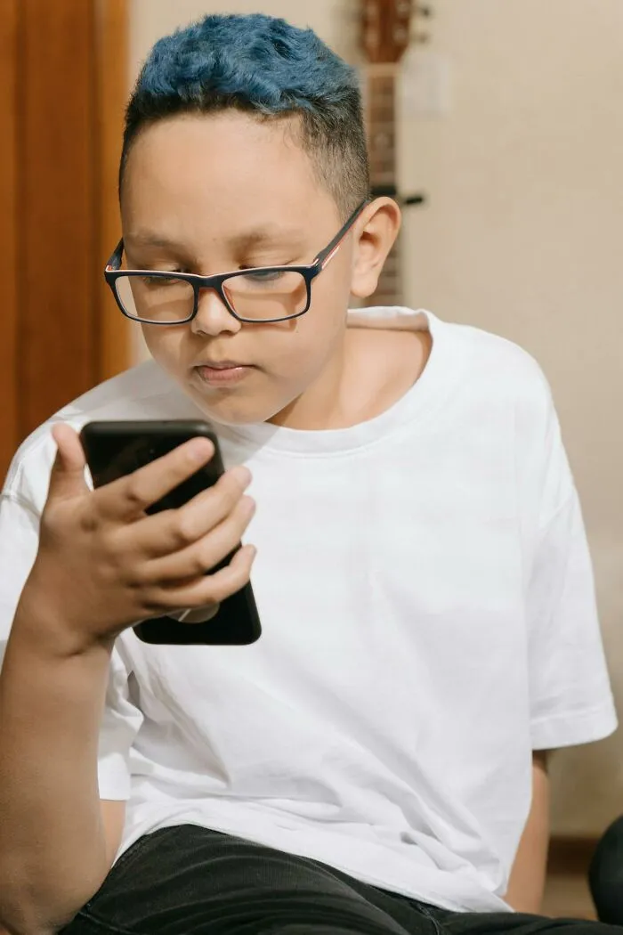 Young boy wearing glasses and a white shirt, looking intently at smartphone, highlighting people silently judge others concept.