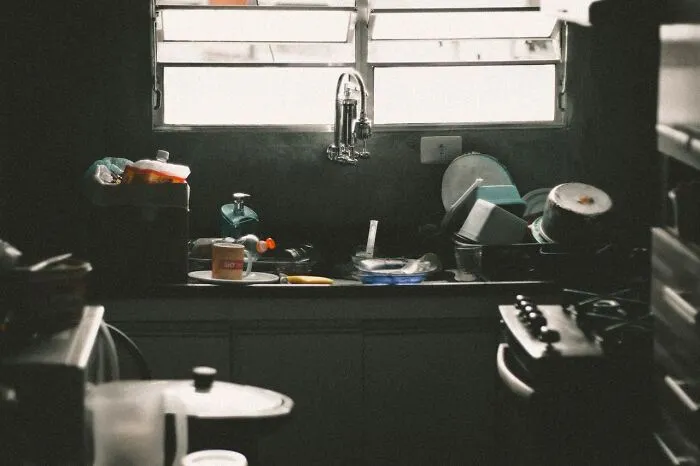 Dimly lit kitchen sink area with dirty dishes overflowing, highlighting neglect and unsafe living conditions related to child safety concerns.