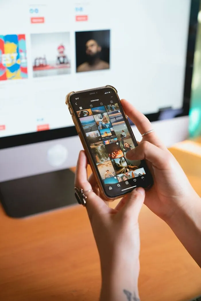 Person silently judging others while scrolling through social media photos on a smartphone at a desk with a computer screen.