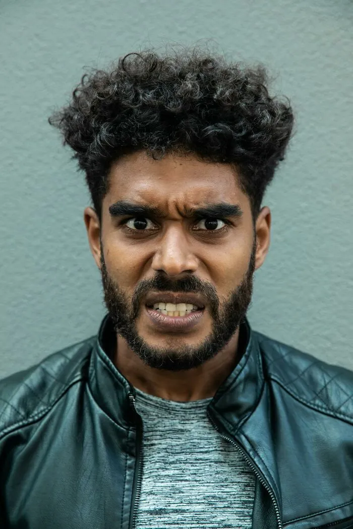 Man with curly hair and beard looking angry and intense, wearing a black leather jacket against a plain background
