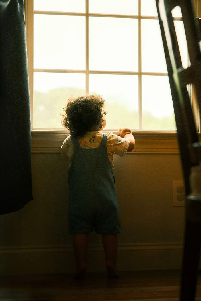 Young child with curly hair standing by a window in a dimly lit room, symbolizing child protection and safety.