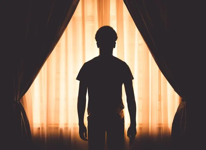Silhouetted figure standing by glowing window curtains, evoking eerie feelings common to graveyard shift workers.