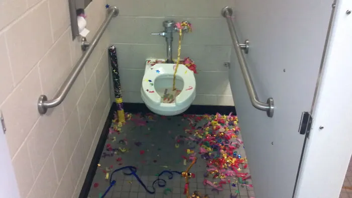 Public restroom toilet with colorful confetti and party streamers scattered on the floor and inside the bowl, funny photo.