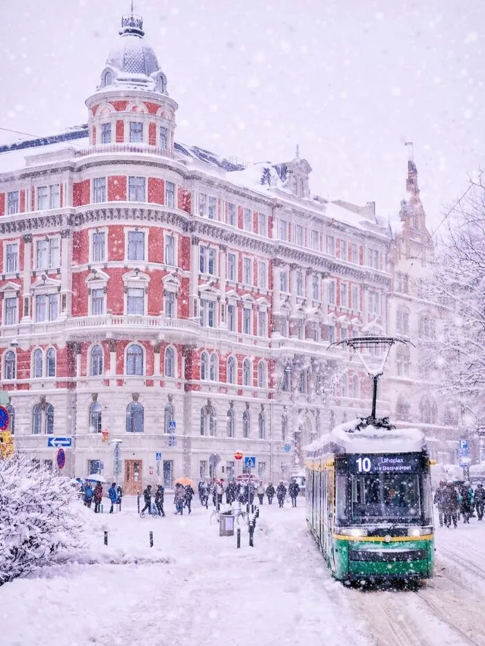 A snow-covered street with a tram and beautiful architecture, featuring a grand red and white building.