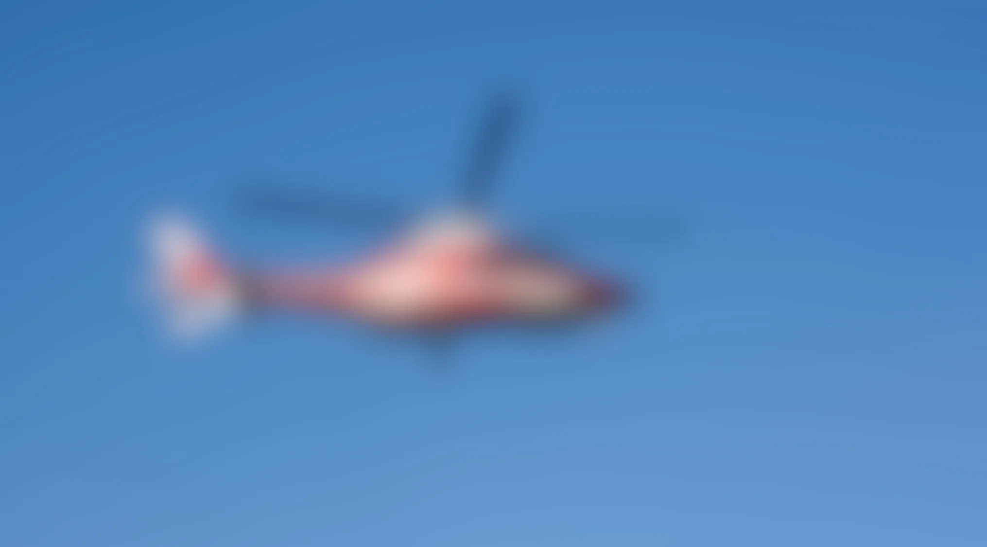 Blurry image of a red and white helicopter flying against a clear blue sky with visible rotor blades.