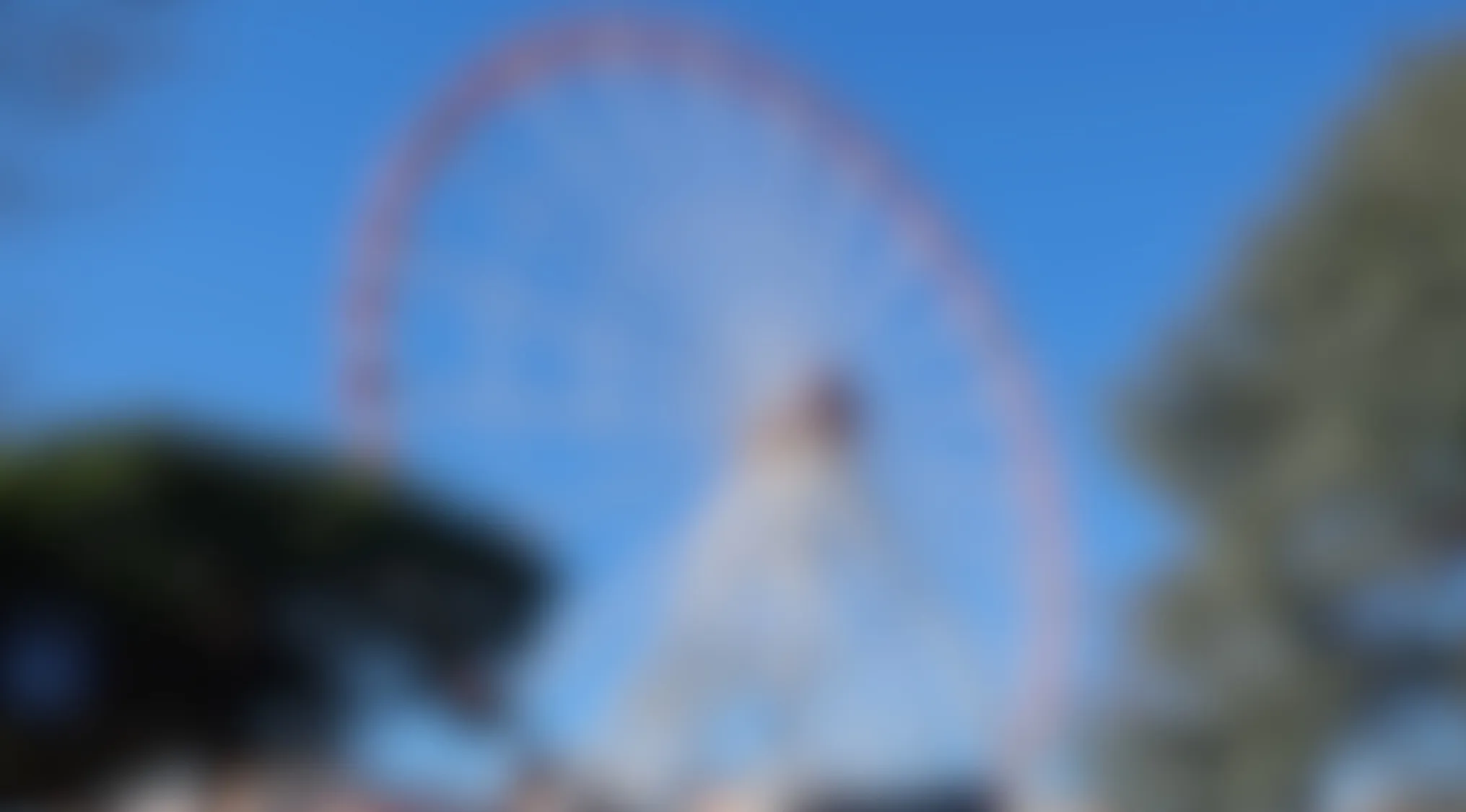 Blurry image of a large Ferris wheel attraction against a clear blue sky with trees nearby