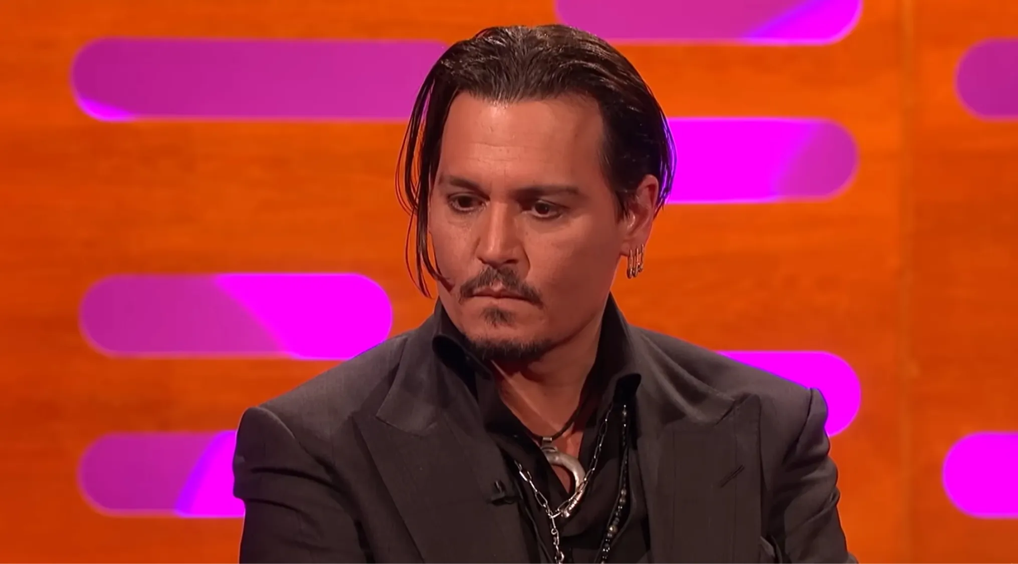 Johnny Depp, famous person, with slicked-back hair and earring in a dark suit on a colorful talk show set