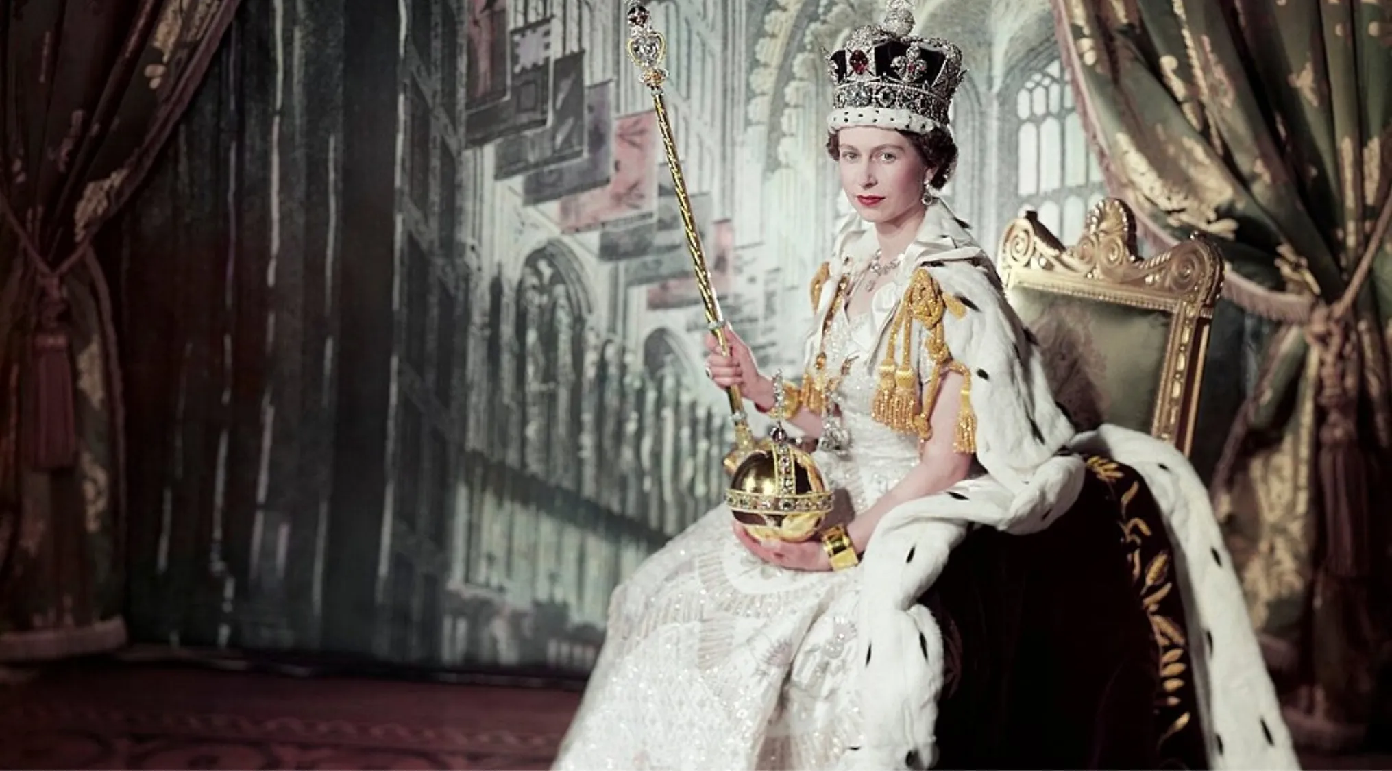 Queen of the United Kingdom in full royal regalia holding orb and scepter while seated on a throne indoors.