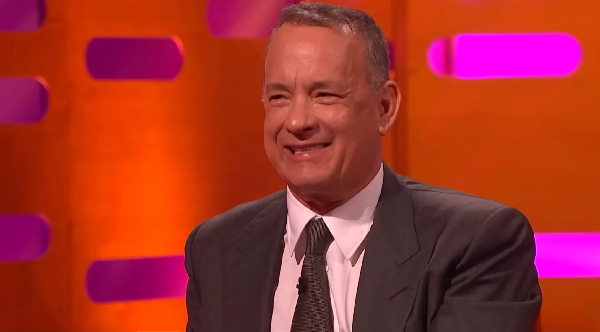 Actor smiling on a colorful studio set, wearing a dark suit and tie, can you identify this famous person?