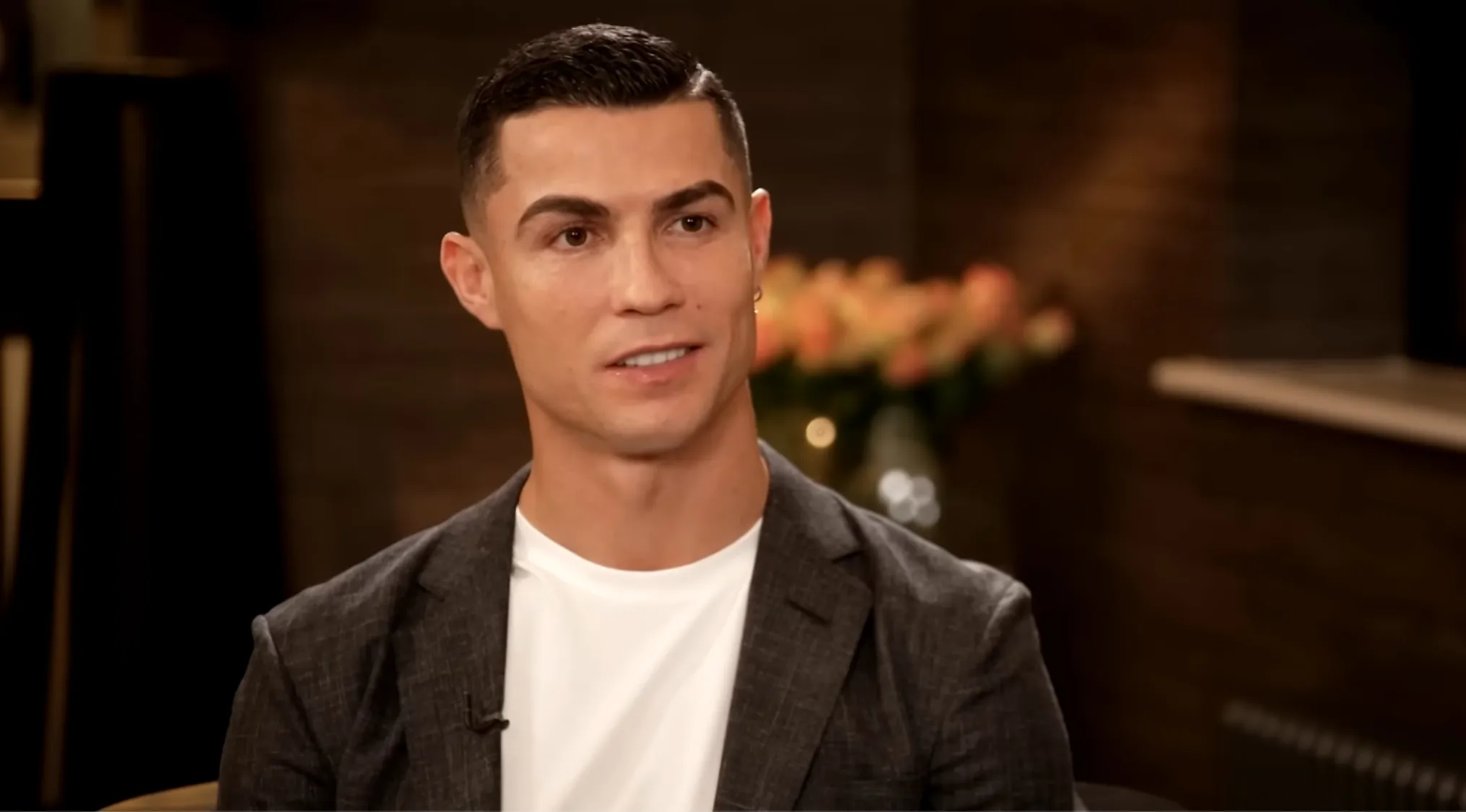 Football superstar in a dark blazer and white shirt, sitting indoors with a blurred floral background.