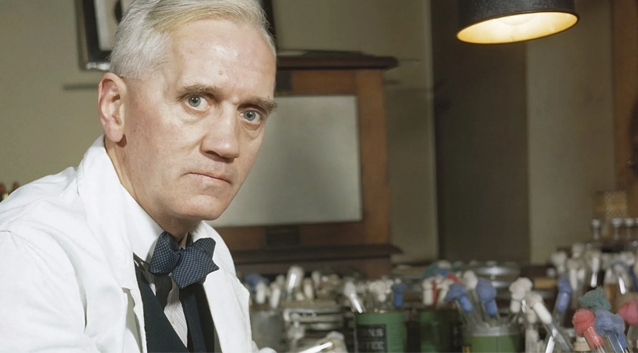 Scientist in a lab coat and bow tie in a laboratory surrounded by scientific equipment identifying discovered penicillin.