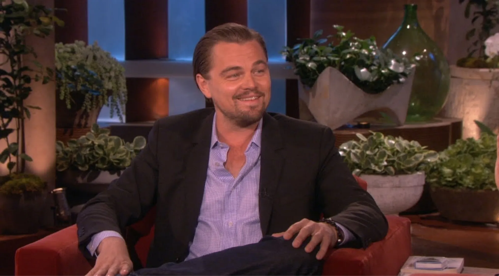 Famous actor smiling during a TV interview, wearing a black blazer and light blue shirt in a studio setting.