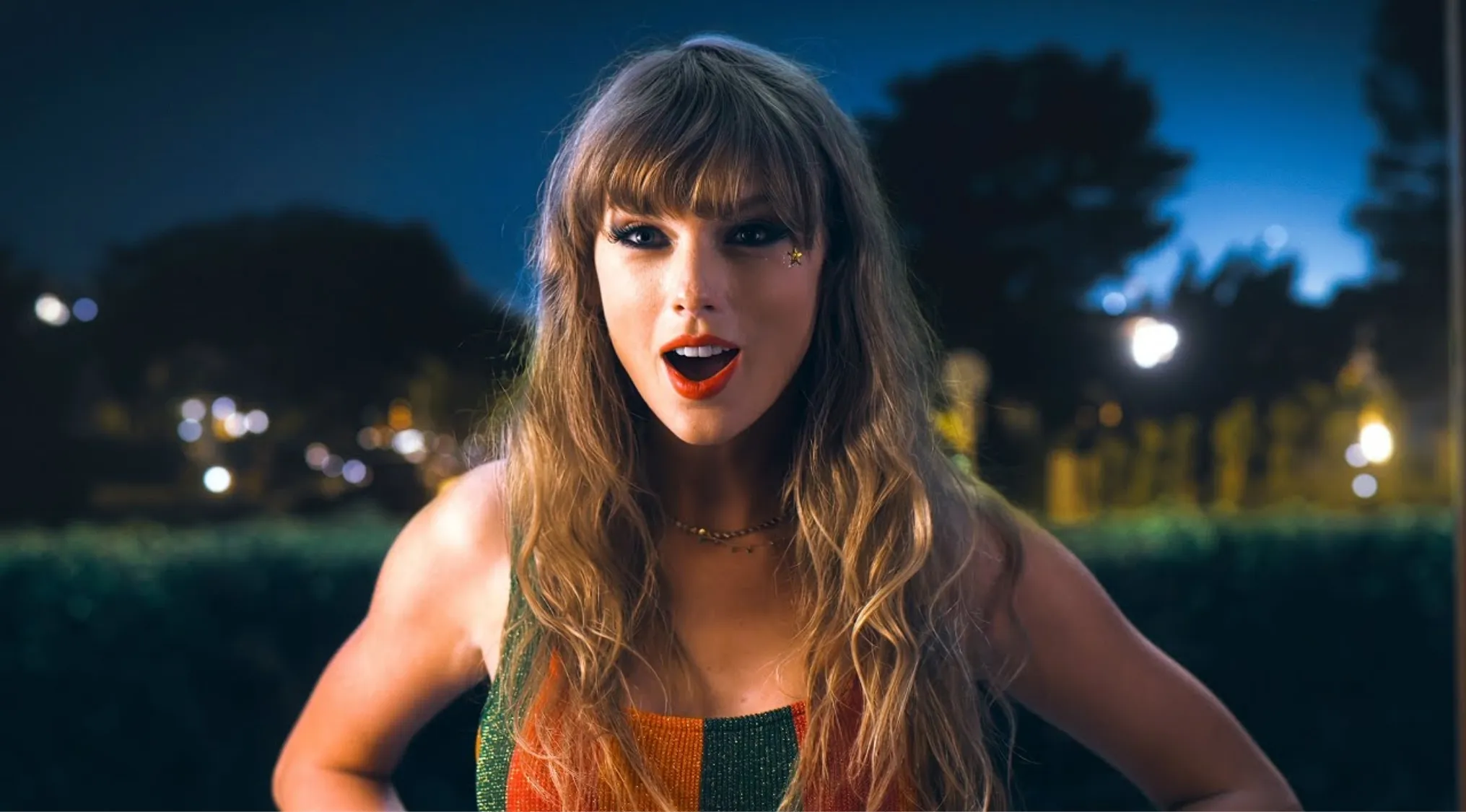 Famous singer posing outdoors at night with long wavy hair and colorful striped top, playful expression.