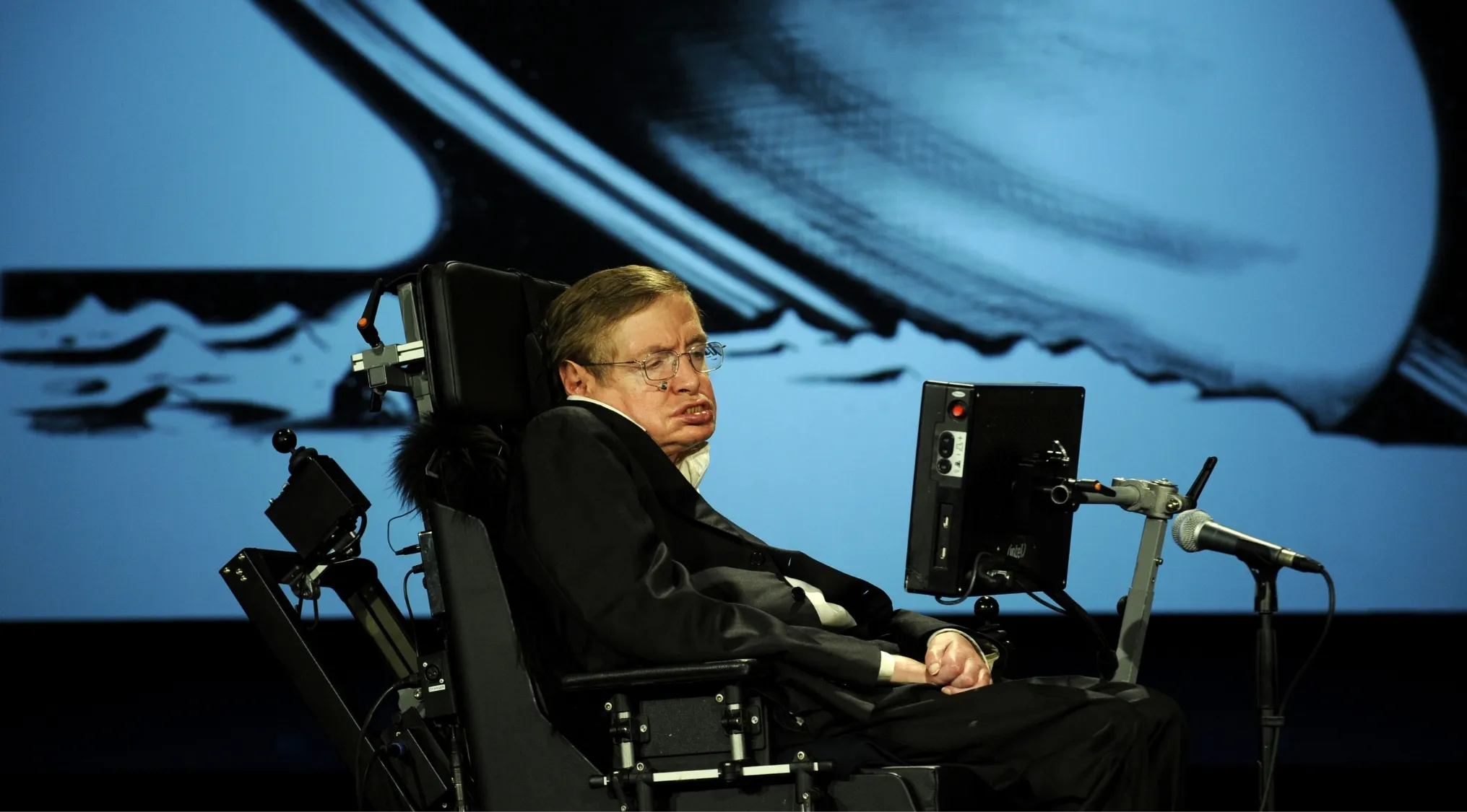 Theoretical physicist known for black holes research speaking at a conference in a wheelchair with a microphone nearby.