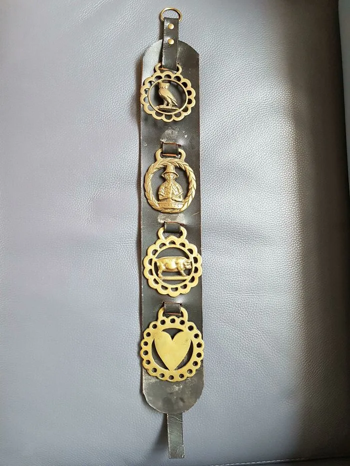 Leather strap with four brass decorative medallions featuring a bird, a soldier figure, an animal, and a heart, unidentified object.