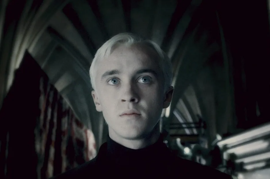 Draco Malfoy with platinum blonde hair, pale skin, and blue eyes standing in a dark, gothic Hogwarts corridor.