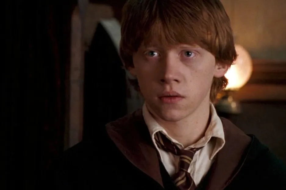 Ron Weasley from the Harry Potter series wearing Hogwarts uniform, with a focused expression in a dimly lit room.