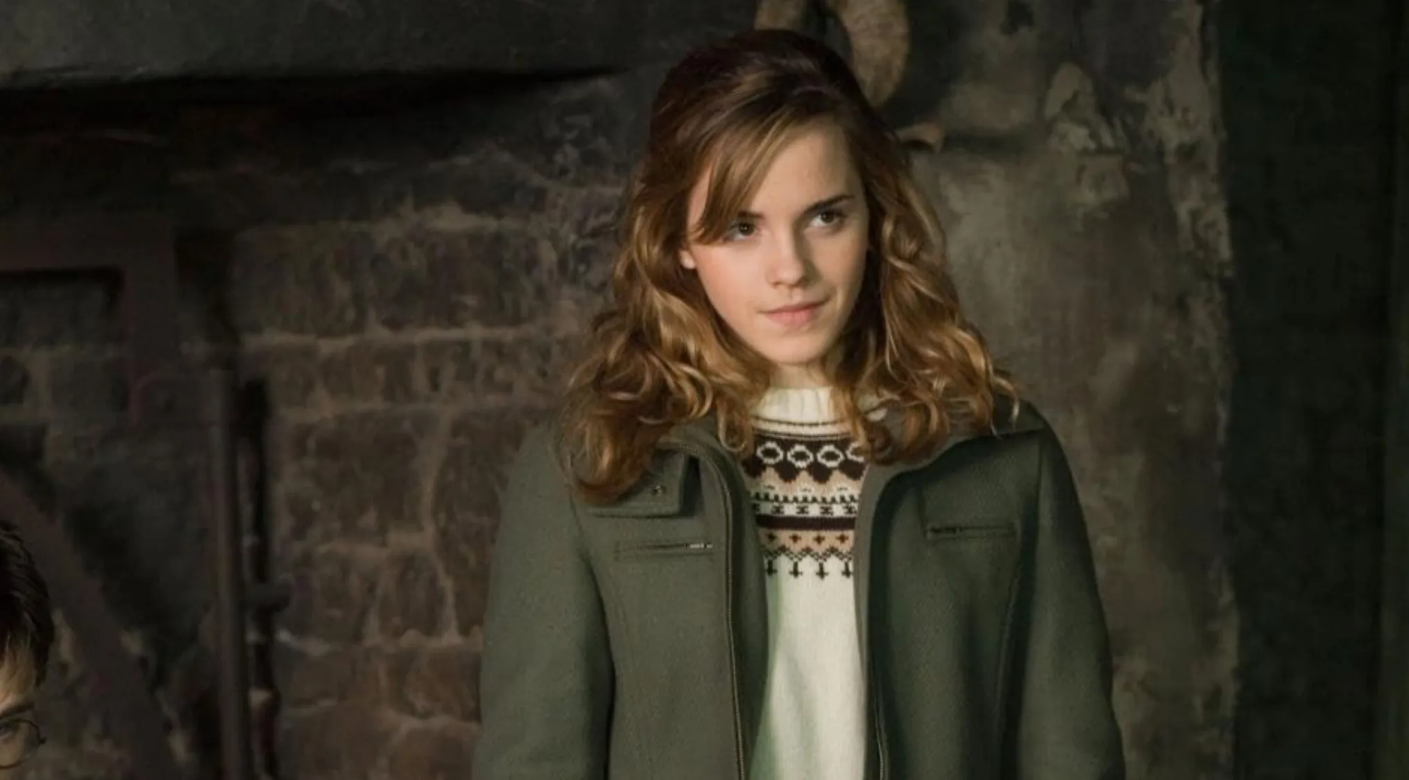 Hermione Granger from Harry Potter series wearing a green coat and patterned sweater standing indoors by stone walls.