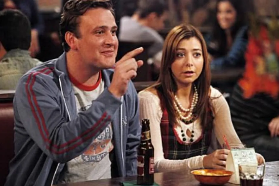 Couple from Friends sitting at a bar table with drinks, engaged in conversation in a casual setting.