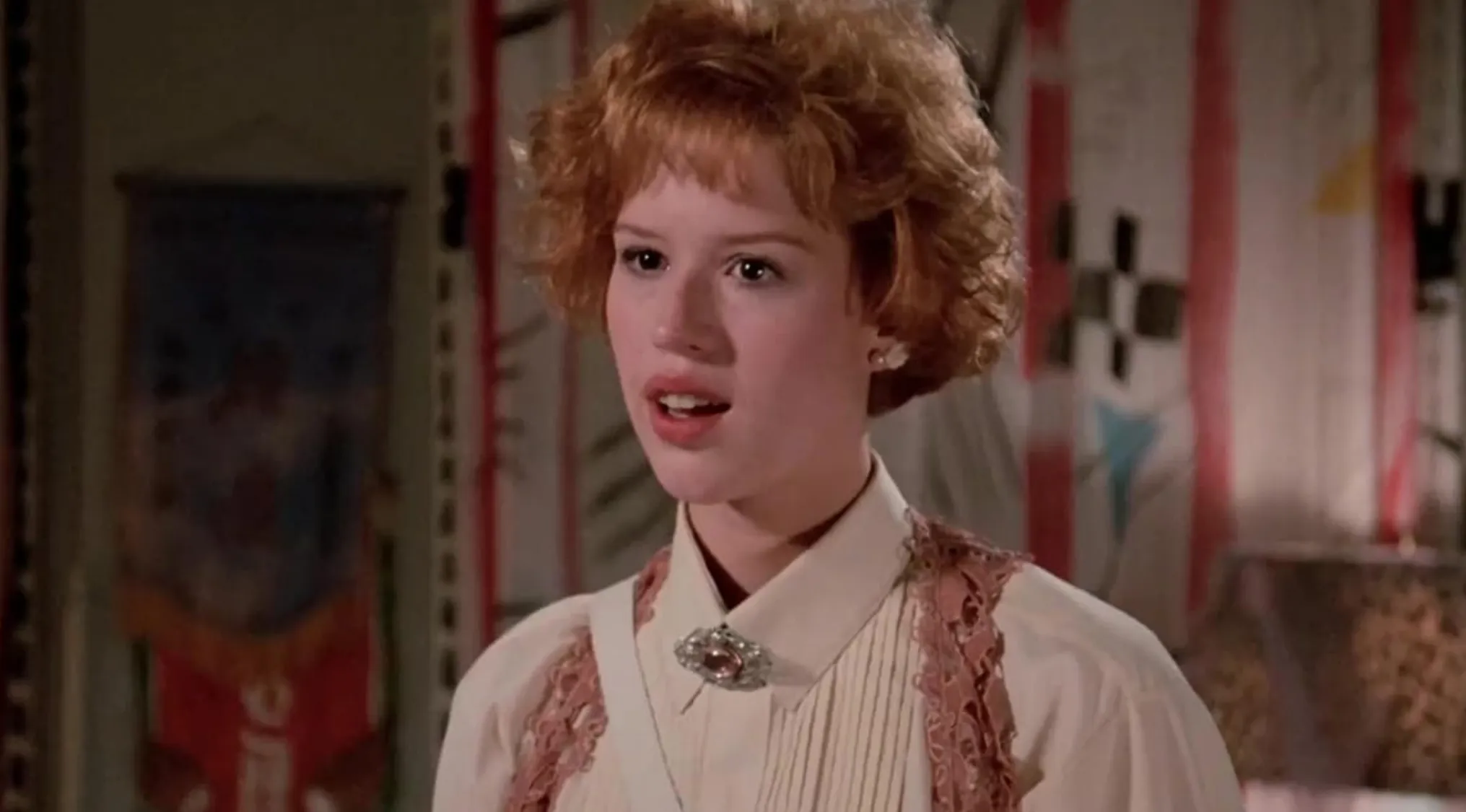 Andie from Pretty in Pink in a vintage blouse and brooch, looking surprised in a colorful room setting.