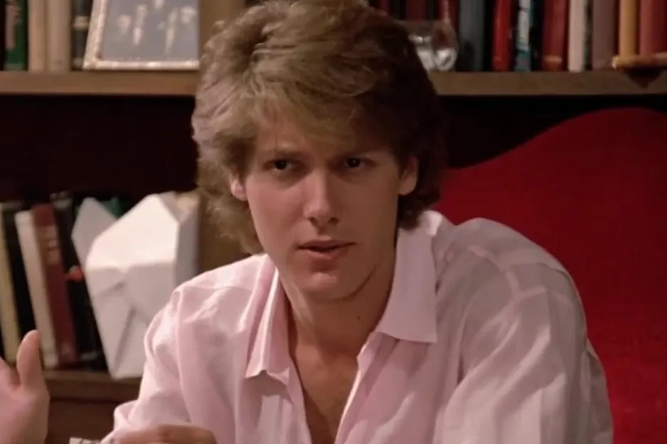 Young man in a pink shirt seated in front of bookshelves, representing a scene related to Andie's romantic choice in Pretty in Pink.