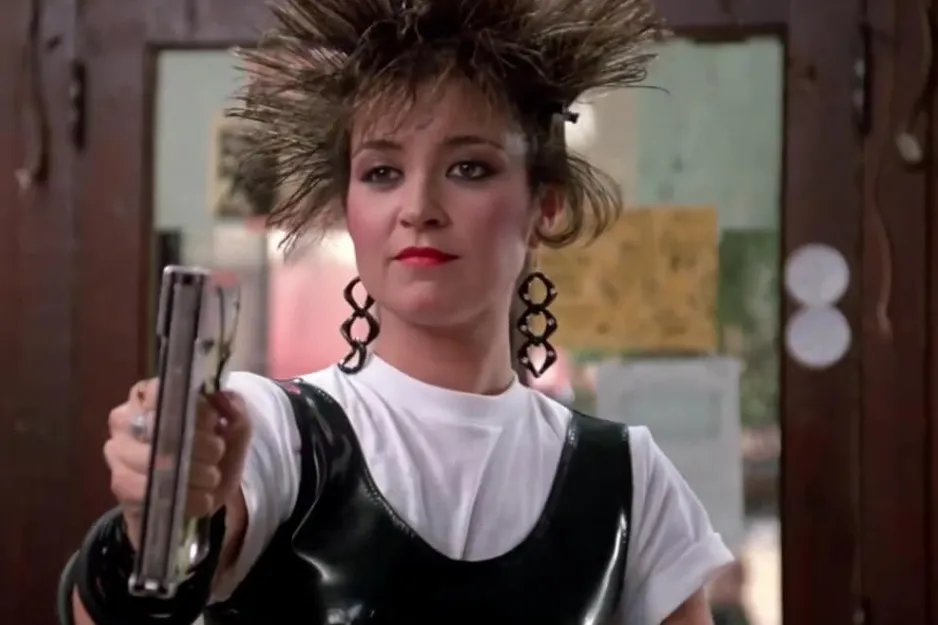Young woman with spiked hair wearing black earrings and a black top over white, holding a gun in a dramatic scene from Pretty in Pink.