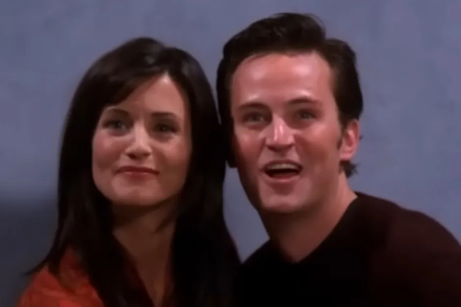Couple from Friends smiling together in a close-up shot with a plain background, showing joyful expressions.