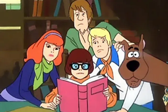 Scooby-Doo characters including Velma, Shaggy, Fred, Daphne, and Scooby reading a large pink book together indoors.