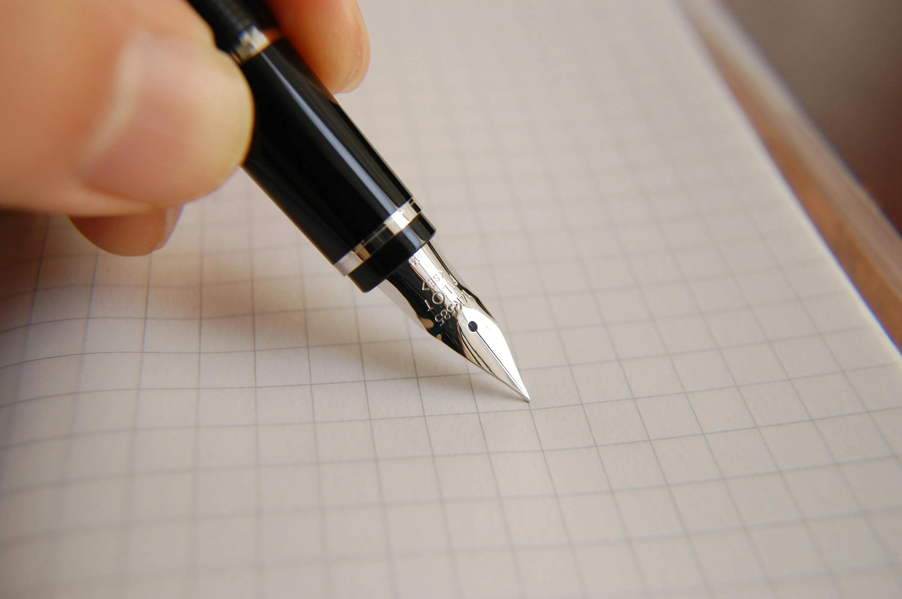 Close-up of a hand holding a black fountain pen poised to write on a sheet of graph paper.