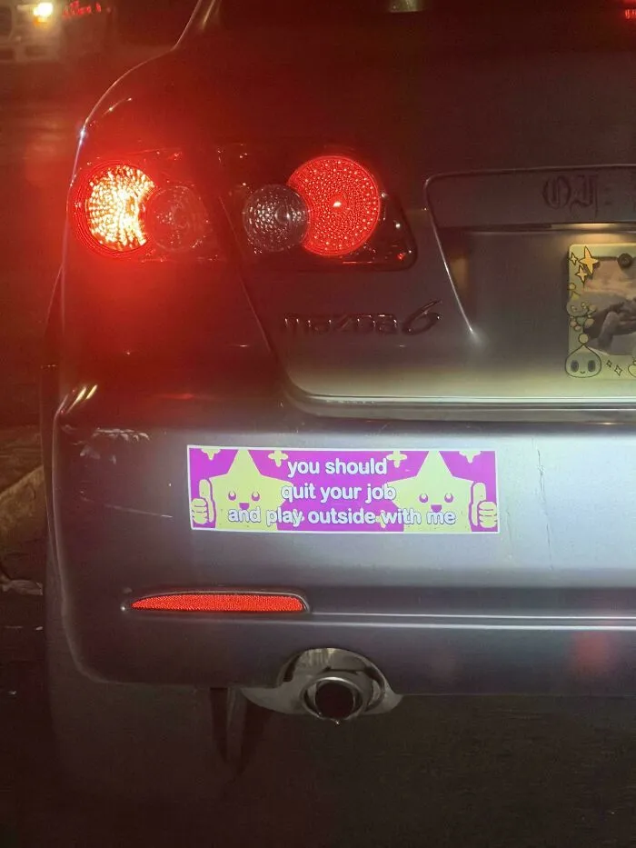 A rear view of a silver car with a funny bumper sticker that reads You should quit your job and play outside with me.