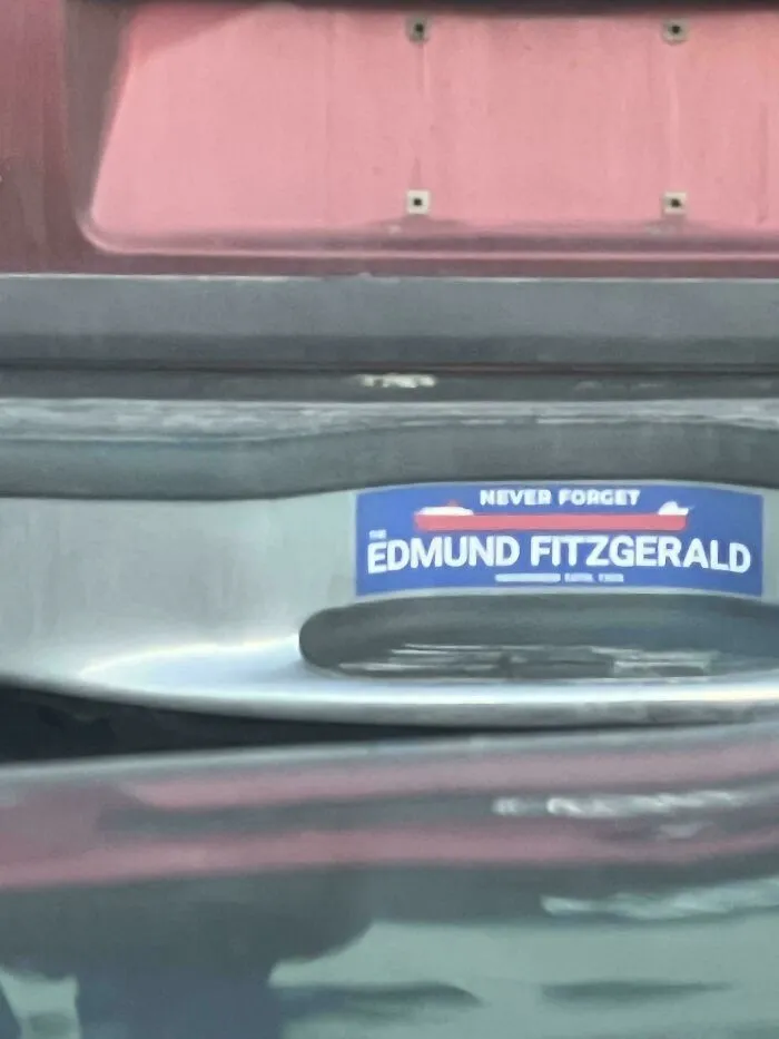 A close-up of a bumper sticker that reads Never Forget Edmund Fitzgerald on a red car, one of many funny bumper stickers.