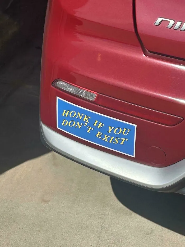 A funny bumper sticker on a red car reads Honk If You Don't Exist in yellow text on a blue background.