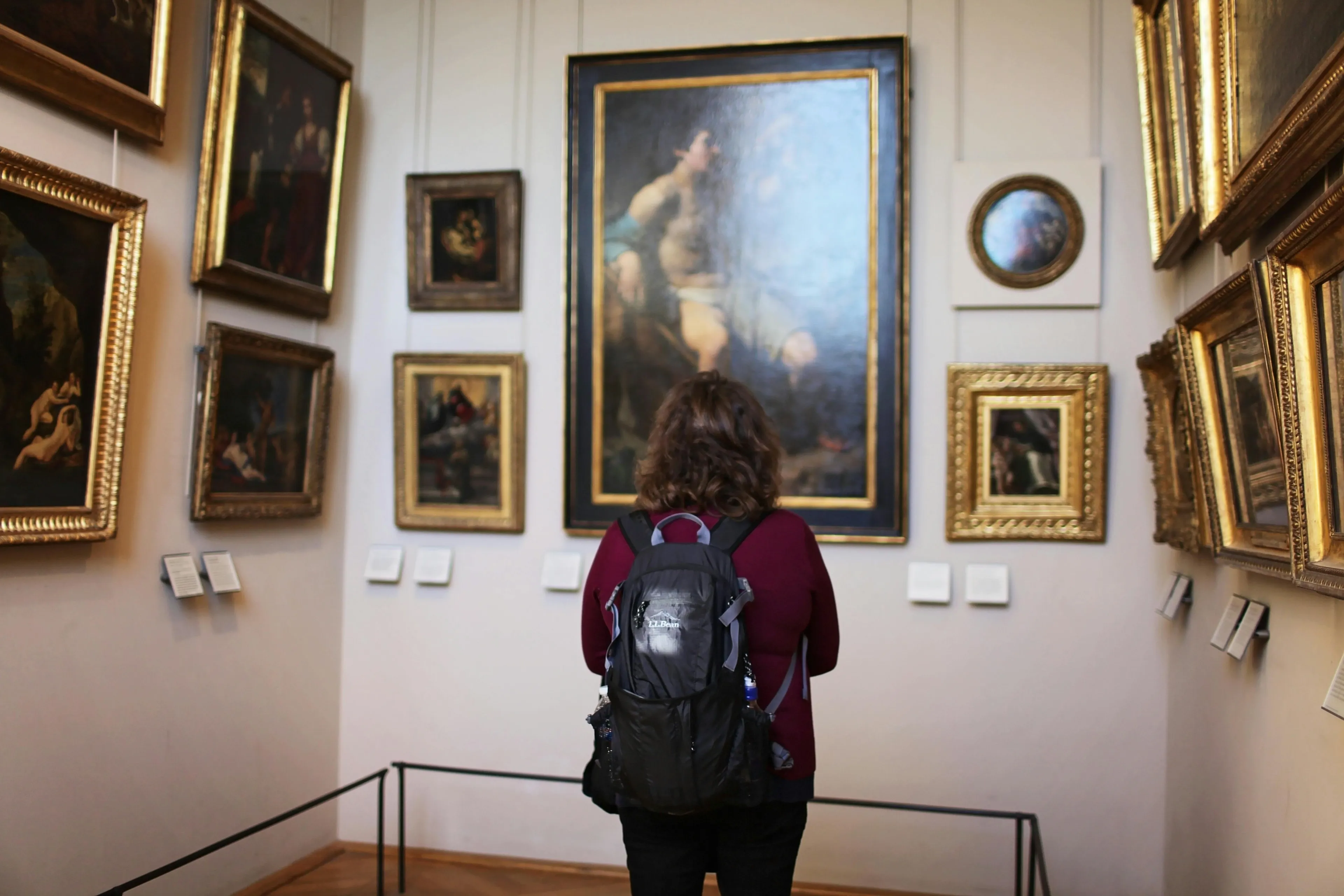 Person with backpack enjoying a solo museum trip, viewing classic framed paintings in a quiet gallery room.