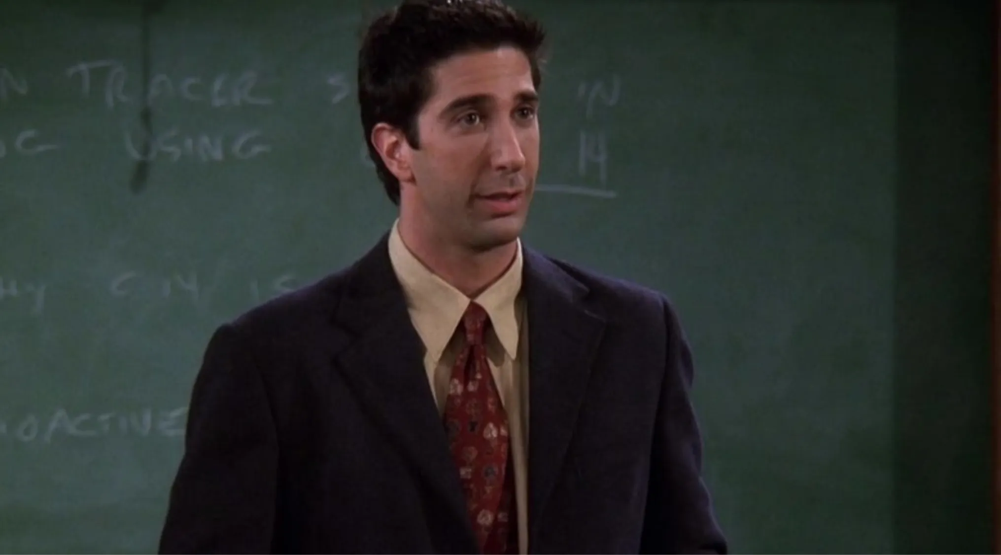 Ross from Friends wearing a suit and tie, standing in front of a chalkboard as a professor in a classroom setting.