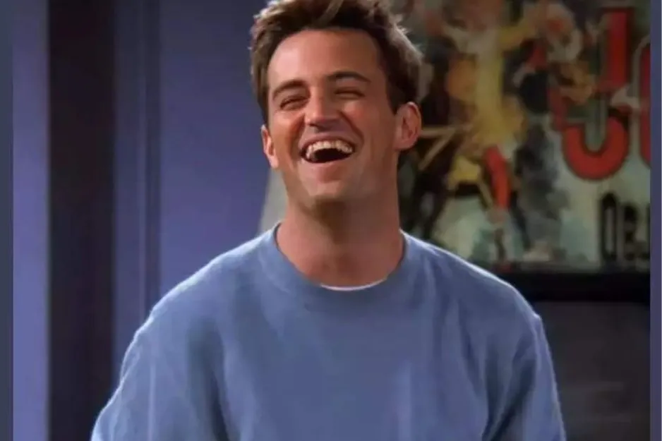 Chandler laughing in a blue sweater with a blurred colorful background in a sitcom setting.