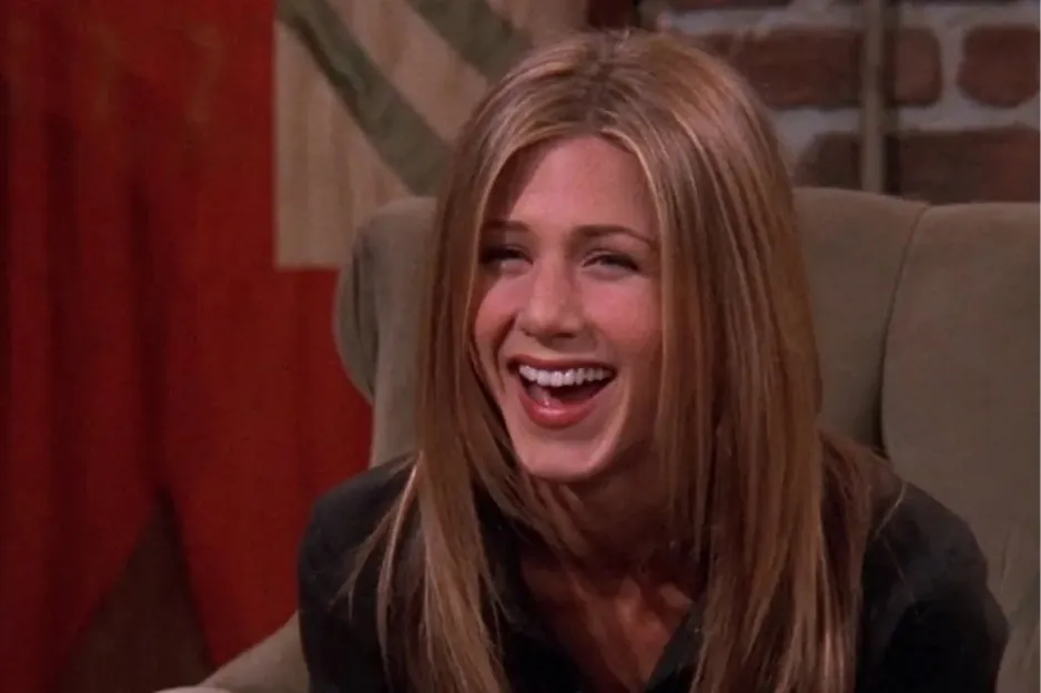 Rachel laughing and sitting on a chair in a cozy indoor setting, smiling brightly in a casual moment.