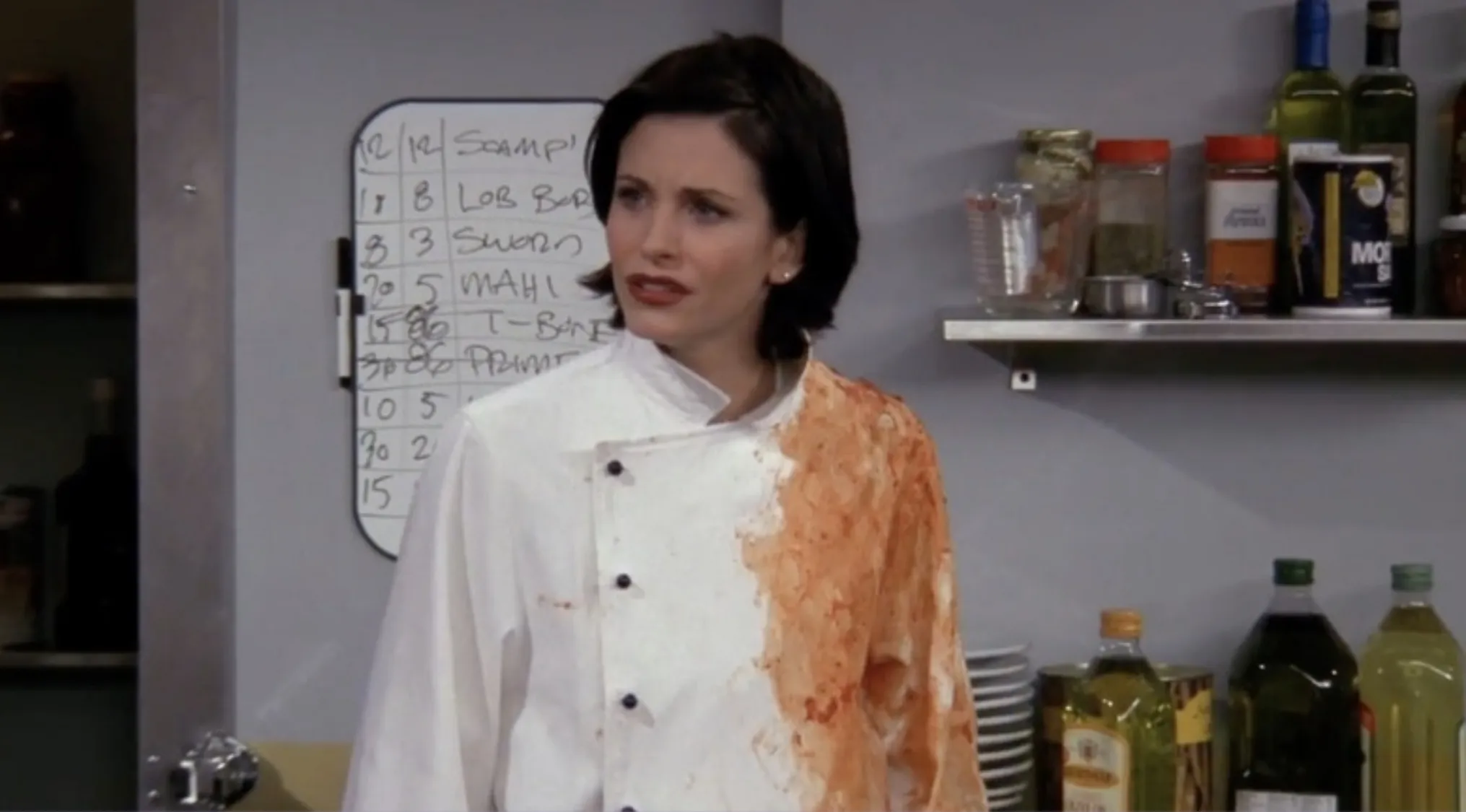 Monica from Friends in a chef coat stained with sauce, standing in a kitchen with cooking ingredients on shelves.