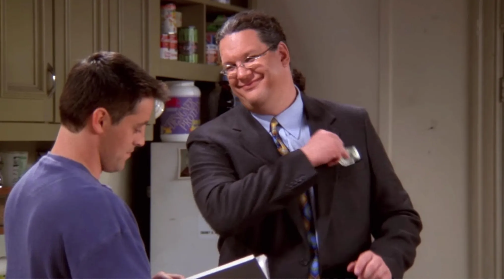 Friends fan moment with Joey holding an encyclopedia volume while a man in a suit smiles in a kitchen setting