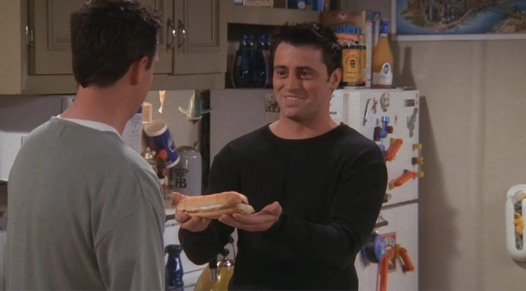 Friends character Joey smiling in kitchen holding a sandwich, proving ultimate Friends fan knowledge about his favorite food.