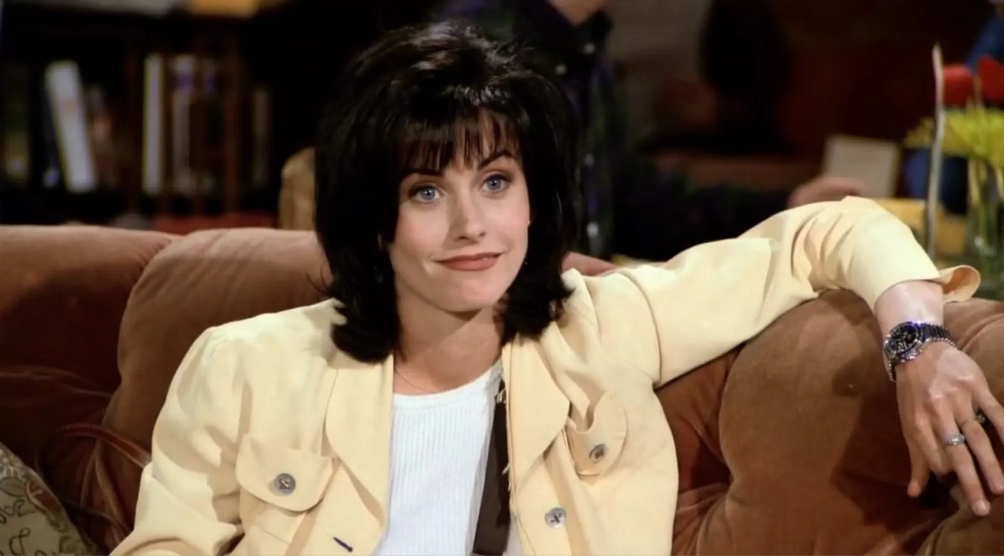 Monica from Friends sitting on a couch with a confident expression, wearing a light-colored jacket and watch.