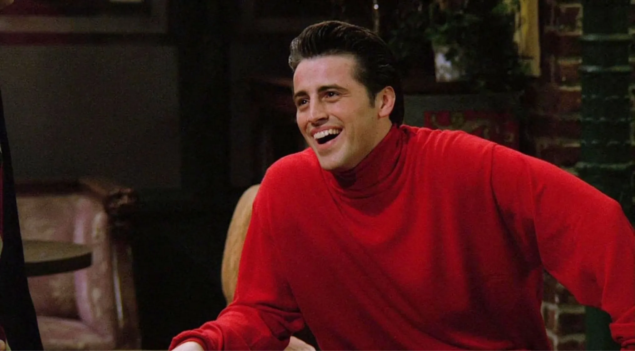 Joey Tribbiani from Friends wearing a red sweater, smiling and laughing in a cozy indoor setting.