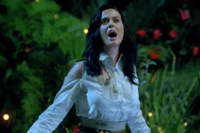 Katy Perry singing Roar outdoors at night surrounded by greenery and colorful flowers in a conflict style themed scene.