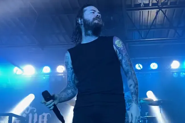 Lead singer of Saliva performing I Walk Alone on stage with blue lighting and visible tattoos on his arms.