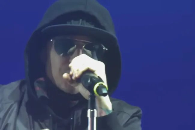Performer wearing sunglasses and a hood singing into a microphone, representing the song Talking To Myself by Linkin Park.