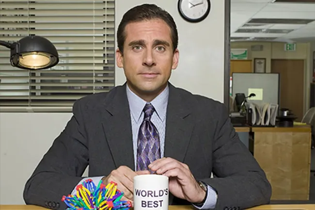 Michael Scott from The Office sitting at his desk with a World's Best Boss mug in a typical office setting.