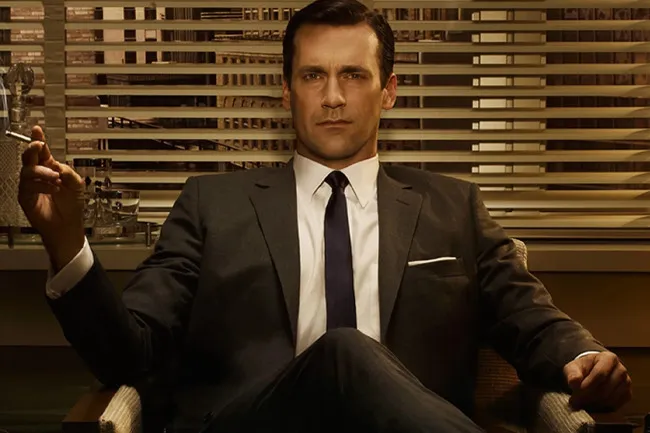 Man in a dark suit sitting confidently, holding a cigarette in an office with blinds behind in Mad Men scene.